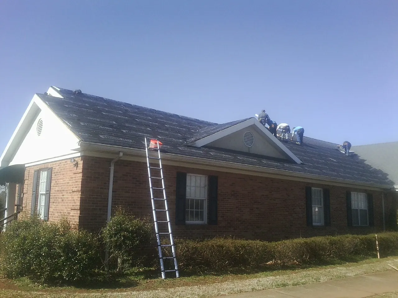 Crew tearing shingles off a brick ranch roof with a ladder set — Emergency Roof Repair work by our crew in Banner Elk