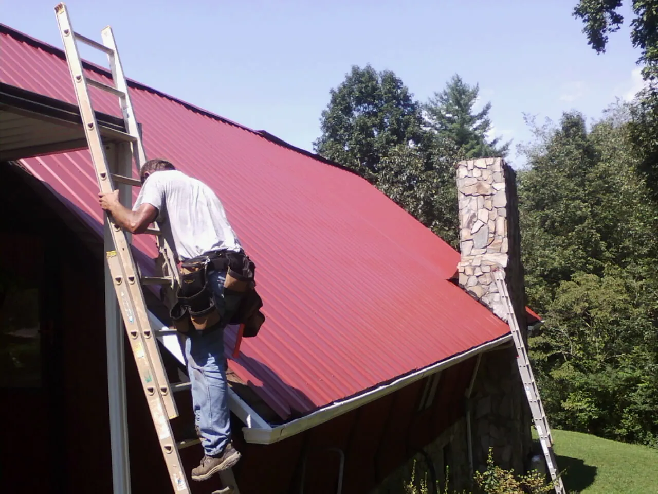 Roofer on a ladder beside a red metal roof and stone chimney — Soffit & Fascia Repair by Roofing Brothers in Banner Elk